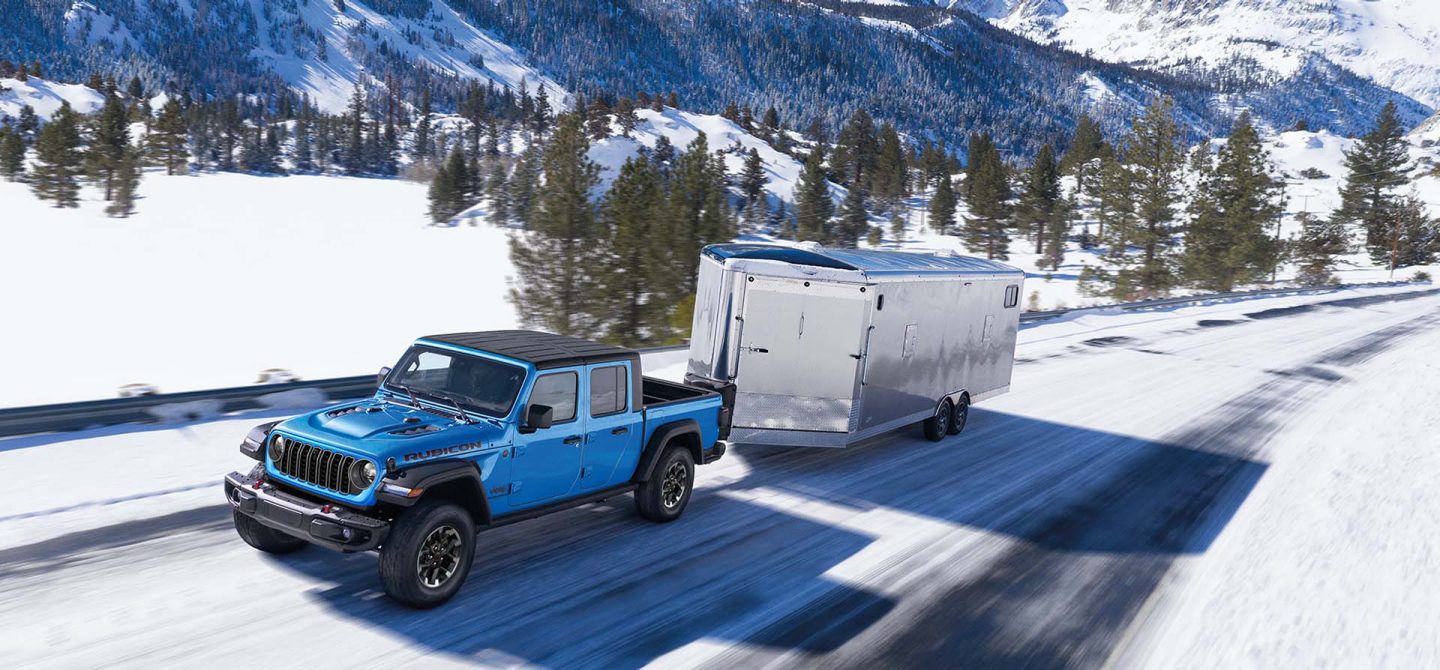 A blue 2025 Jeep Gladiator Rubicon being driven on a snowy highway as it tows an enclosed utility trailer.